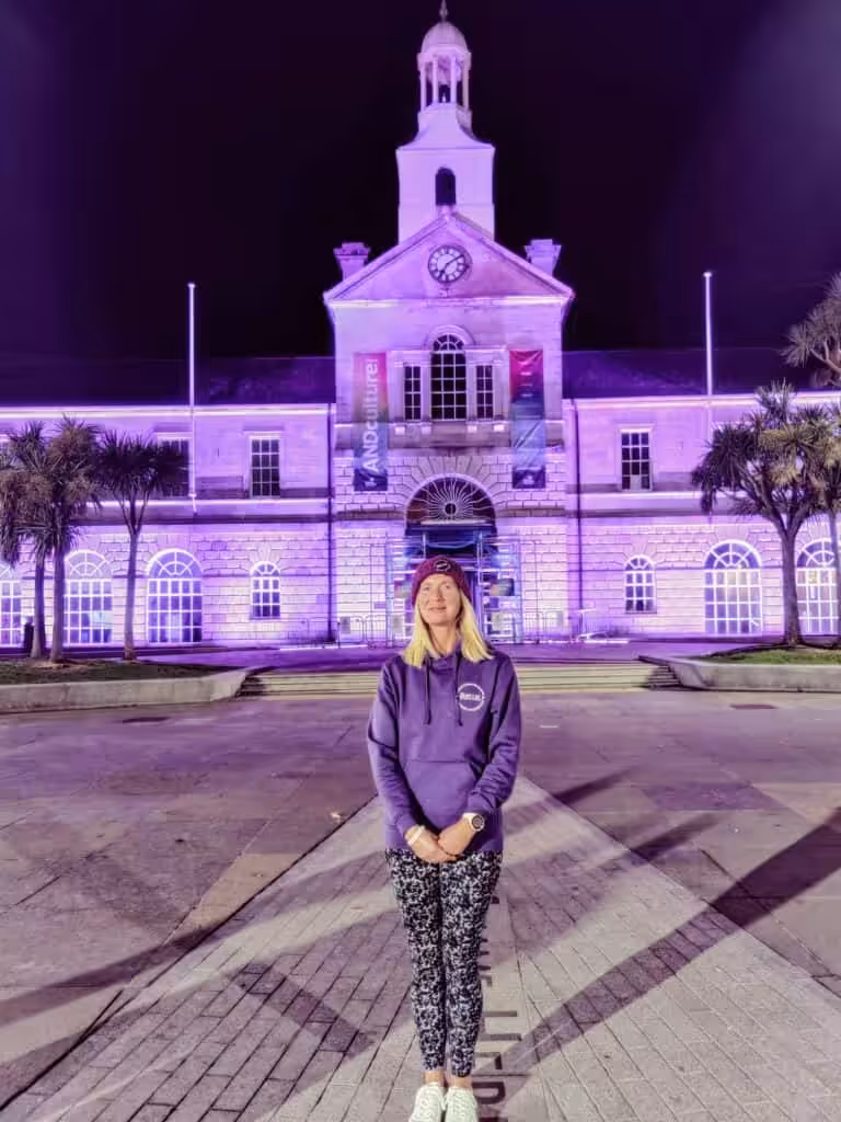 Woman in a purple pots uk hoodie, in front of a building with a clock tower lit up in purple